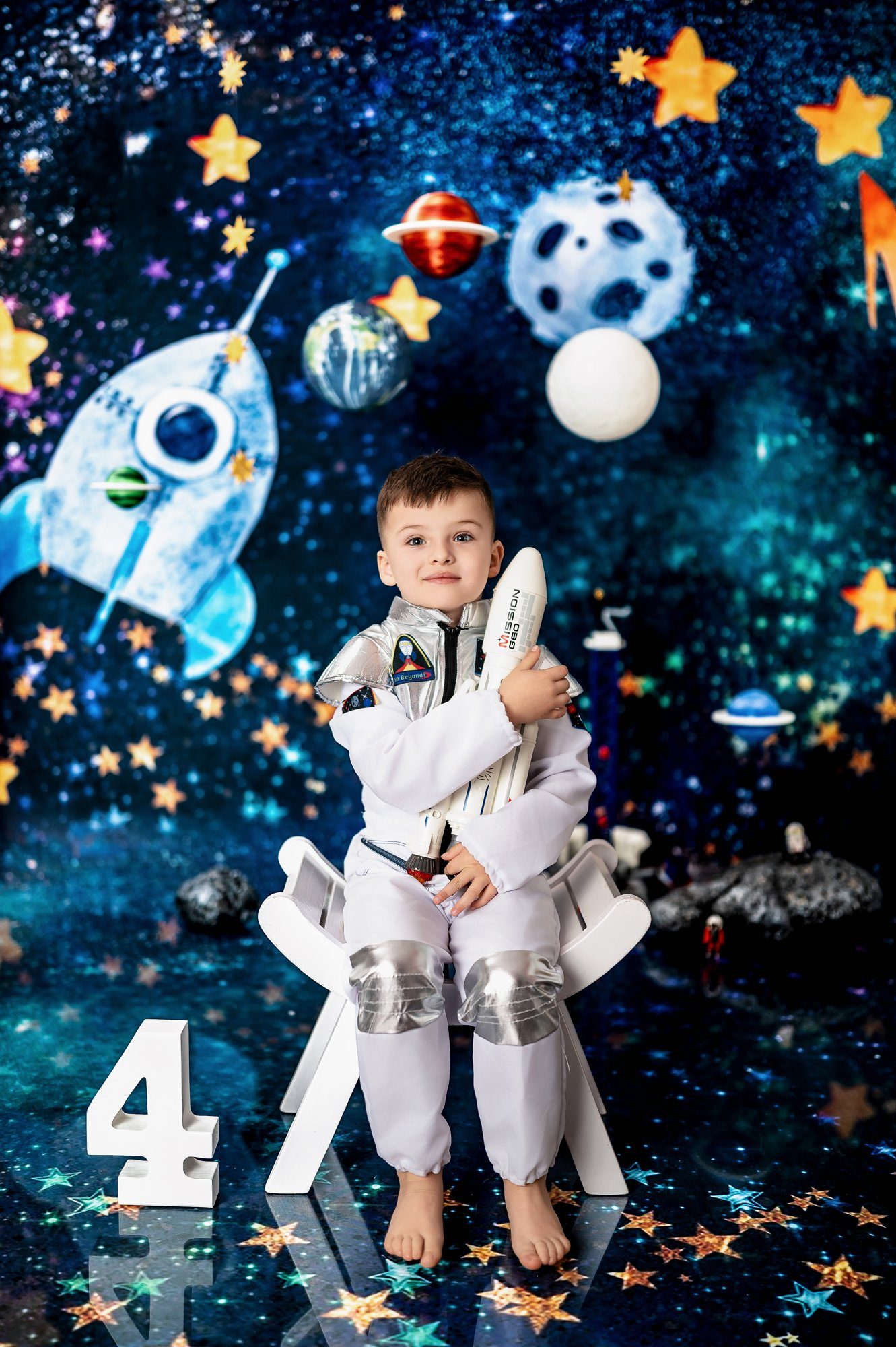 Child in white astronaut costume holding toy rocket with number 4, sitting against sparkling space scene with planets and stars