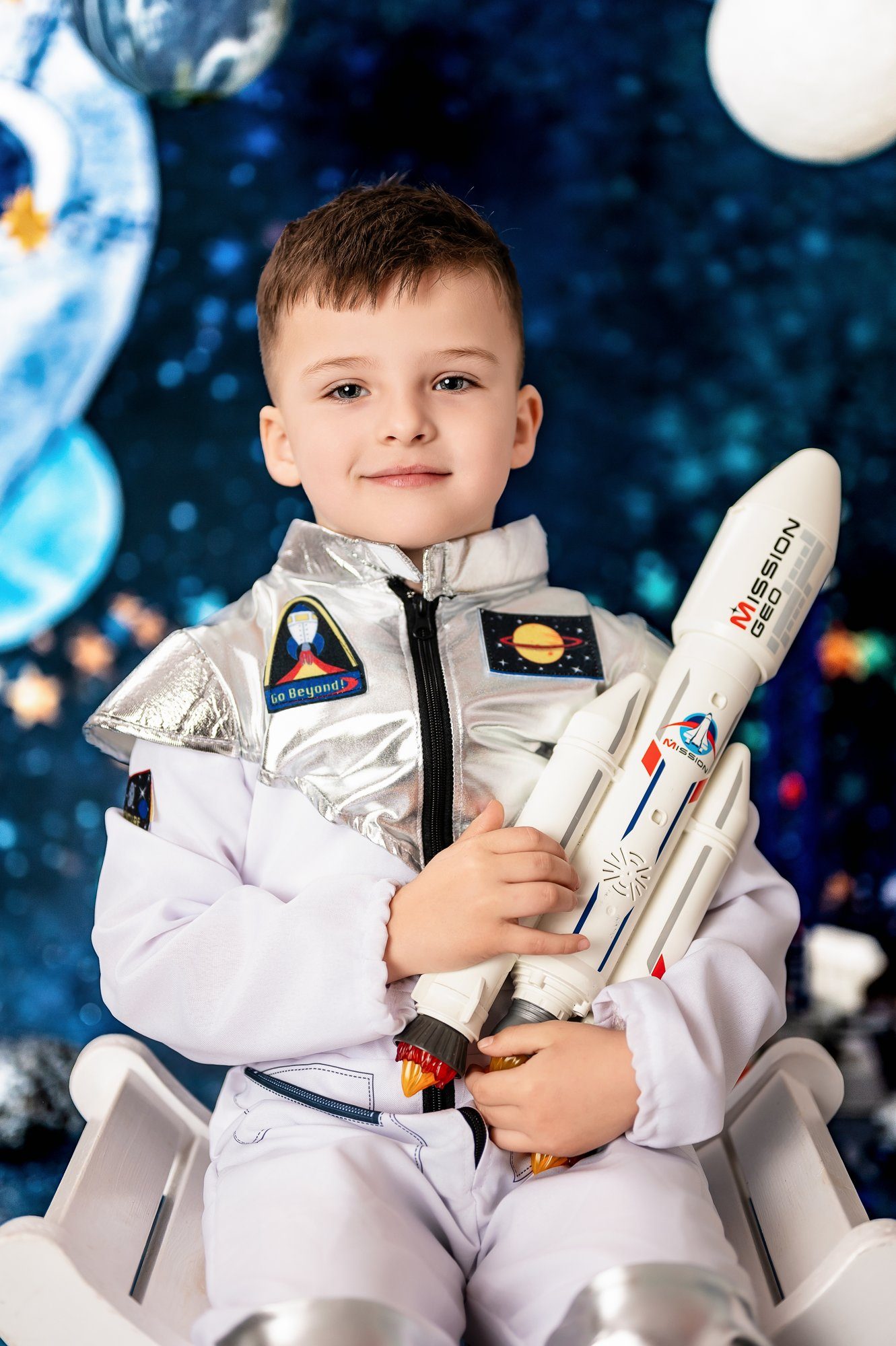 Young boy in light purple astronaut suit holding white toy rocket, smiling against blue cosmic backdrop with planets