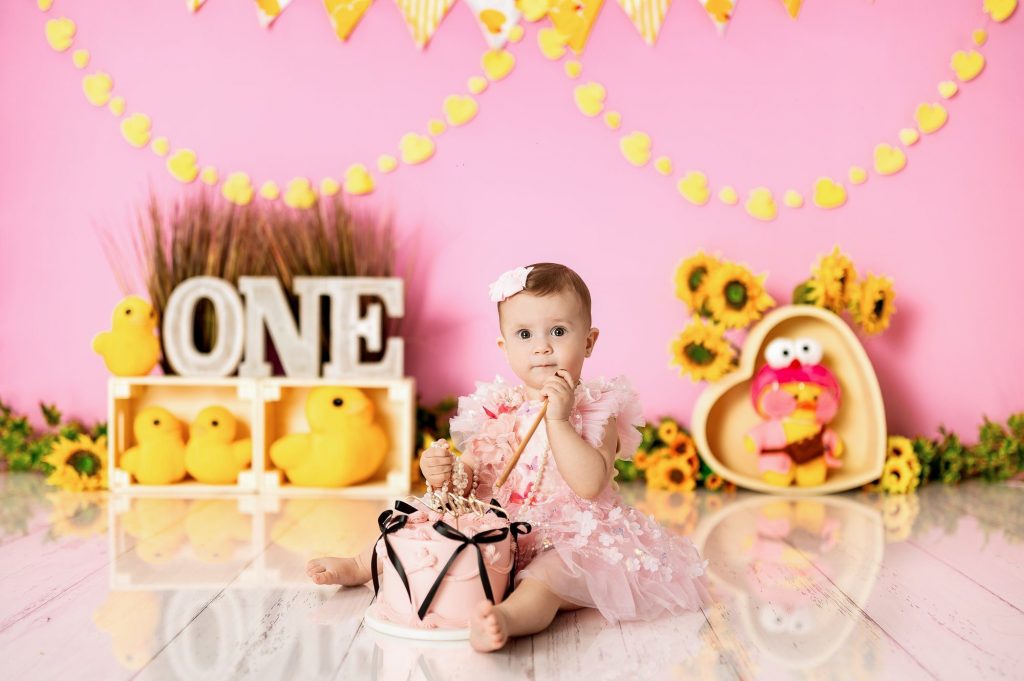 One year old baby girl in pink tutu smashing cake with yellow decorations and sunflowers at Milton Keynes cake smash studio