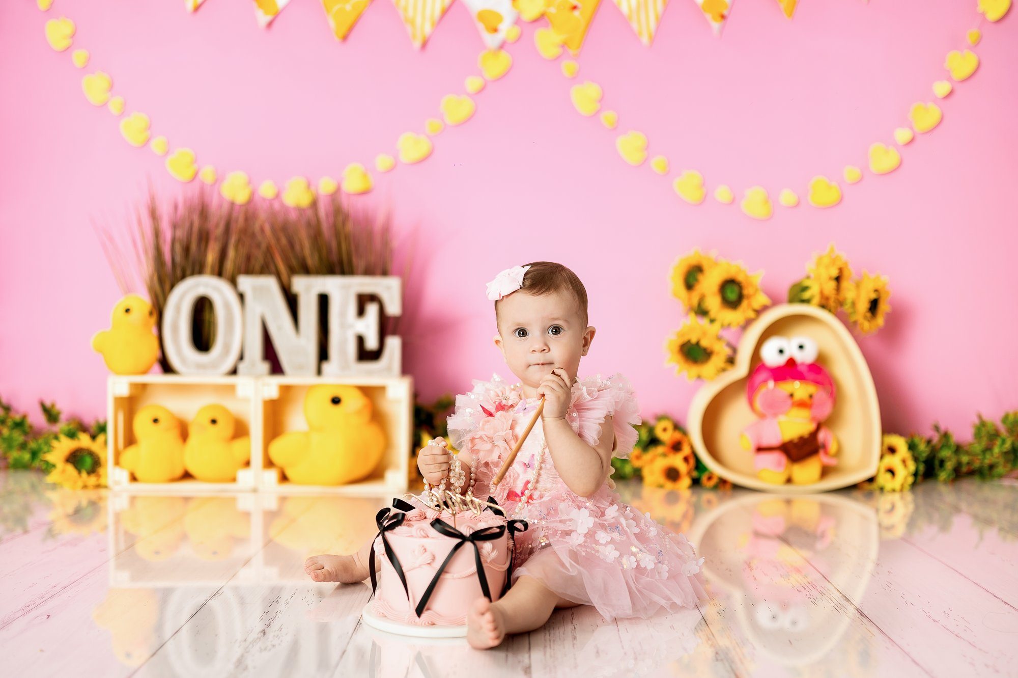 One year old baby girl in pink tutu smashing cake with yellow decorations and sunflowers at Milton Keynes cake smash studio