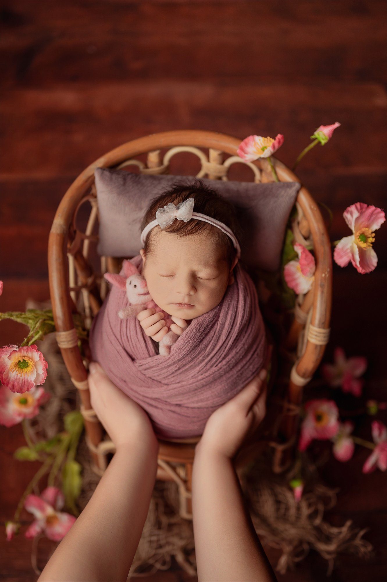 Sleeping newborn baby wrapped in mauve fabric held in parent's hands with pink flowers background