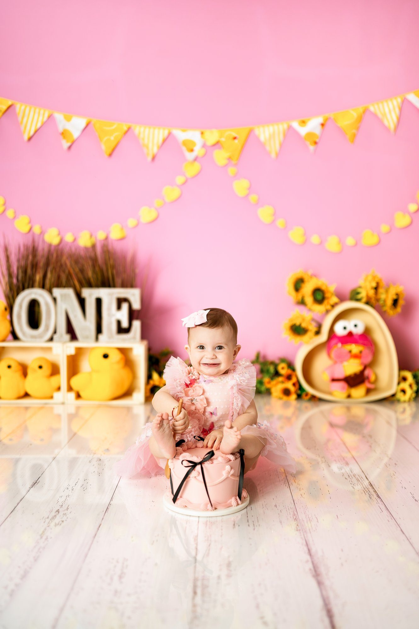Baby girl in pink dress standing on white chair next to number one during first birthday cake smash photoshoot