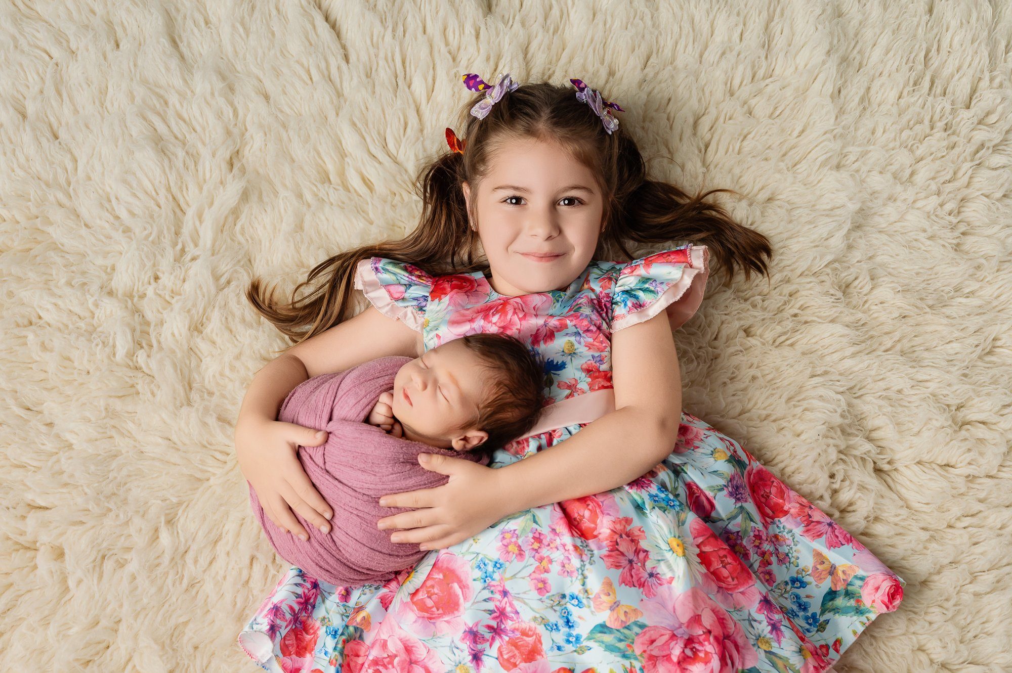 Big sister holding newborn baby wrapped in pink on cream fur backdrop at Milton Keynes studio