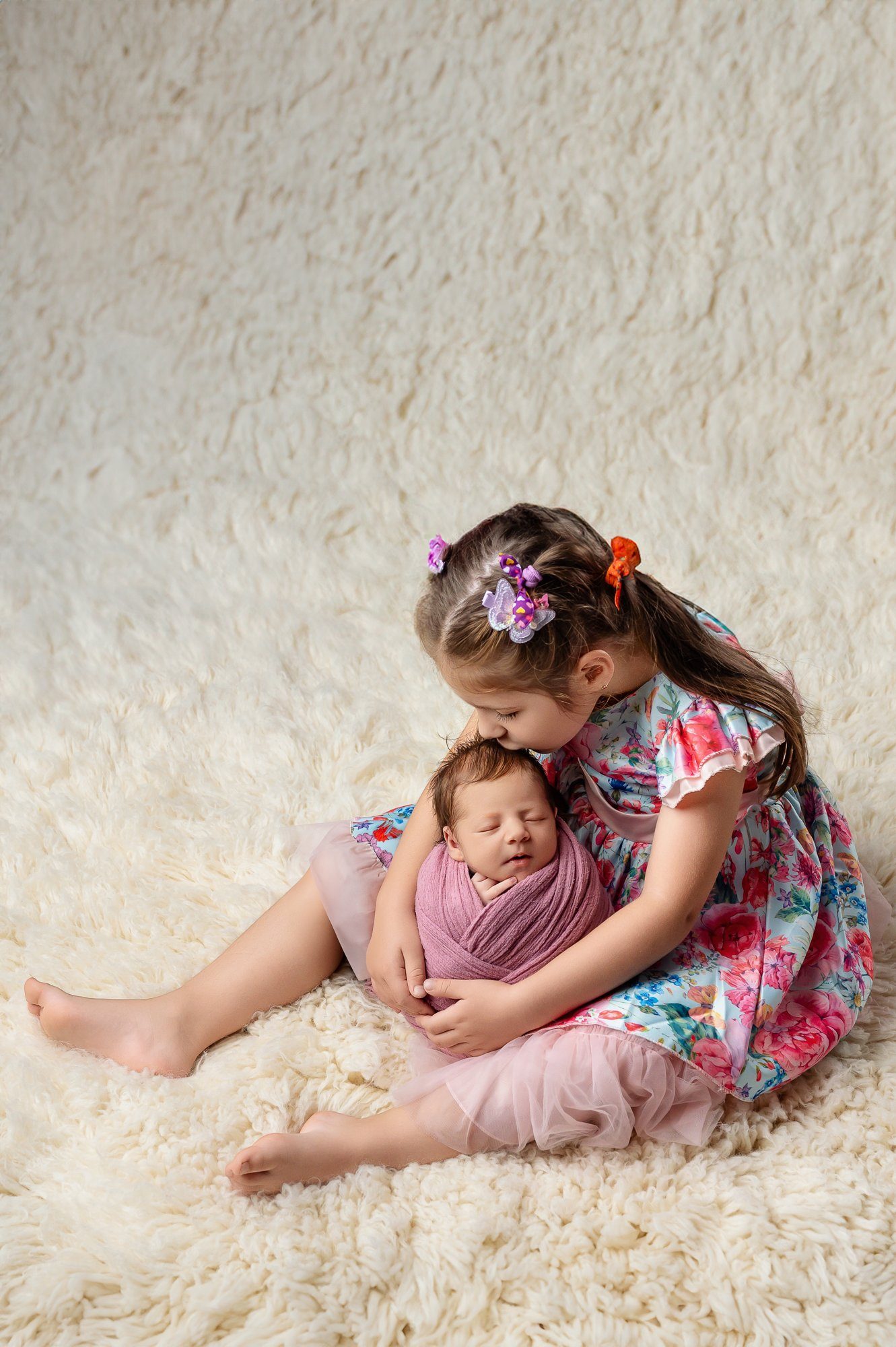 Older sister gently holding and kissing sleeping newborn wrapped in pink fabric on cream fur