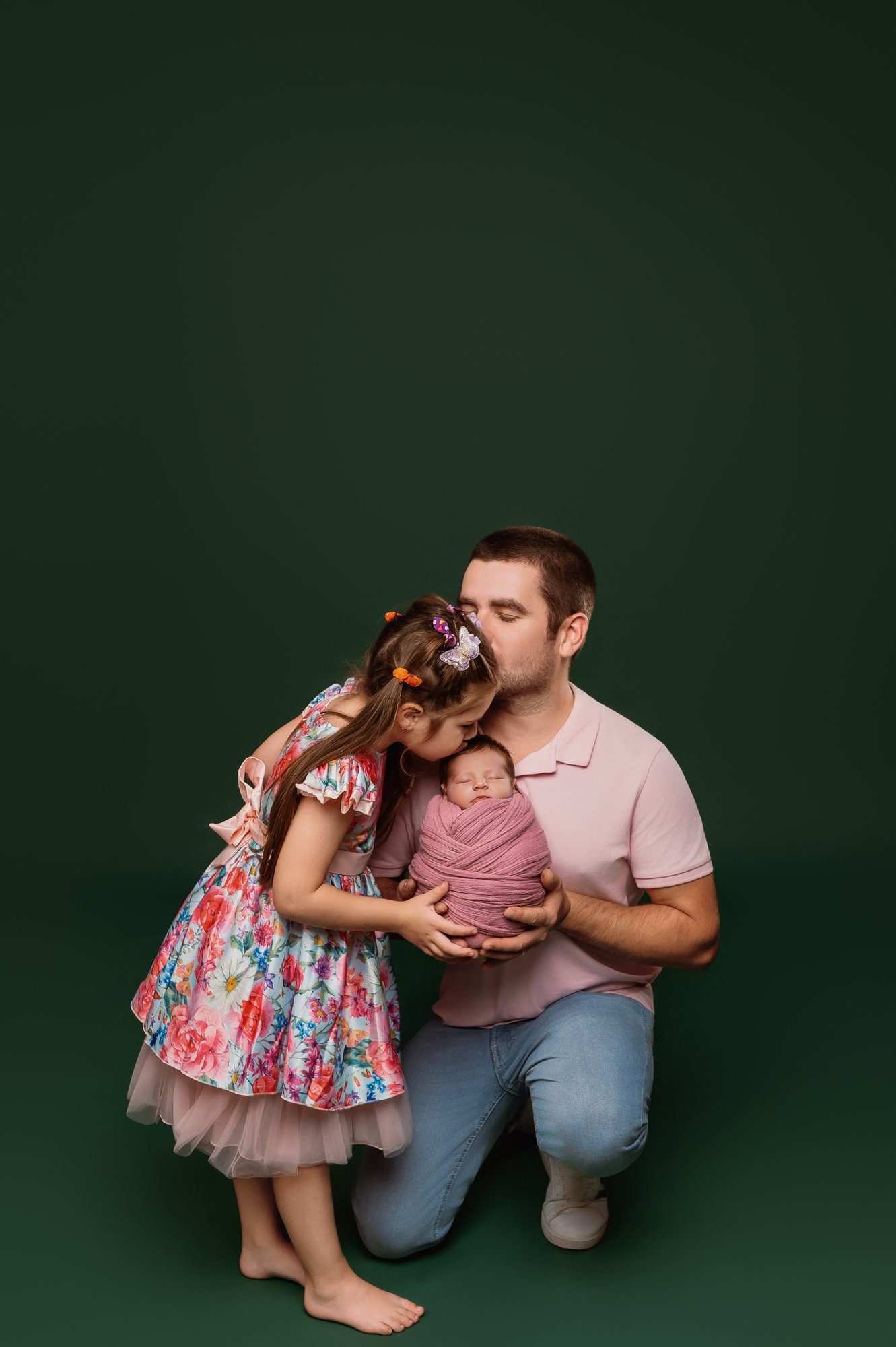 Father holding newborn with big sister in floral dress against green studio backdrop