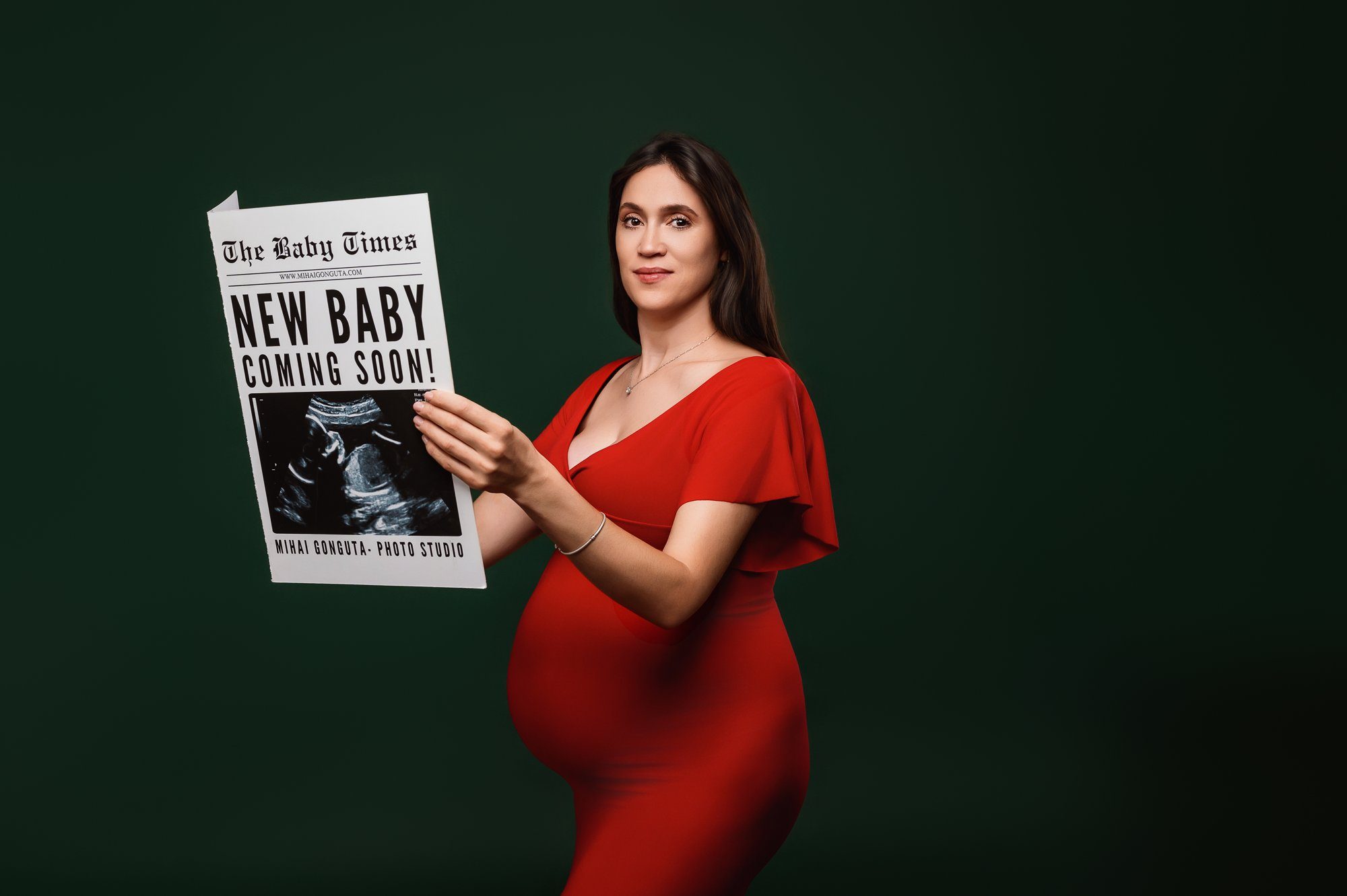 Pregnant woman holding Baby Times newspaper announcing new baby, wearing red dress against green background