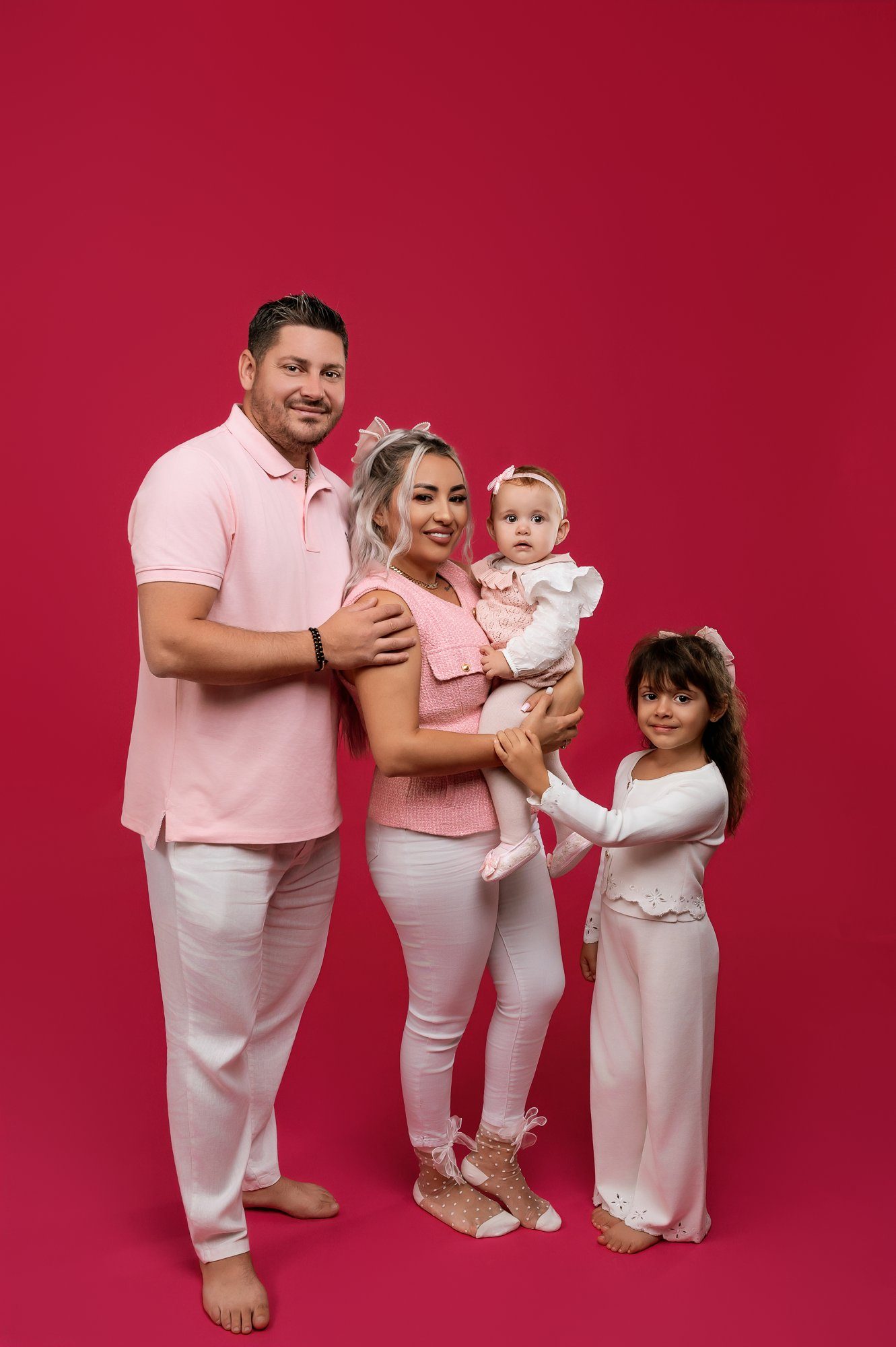 Family portrait of parents and two children wearing coordinated pink outfits against vibrant red studio backdrop