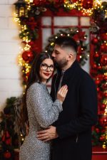 Couple embracing by a red door framed with lights and garlands, Christmas photoshoot