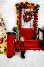 Baby in a red tartan outfit beside a red postbox and a wreath door in the snow