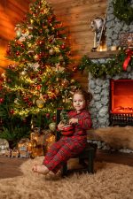 Girl in red pyjamas on a stool beside a Christmas tree and stone fireplace