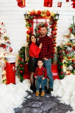 family of three mum dad and young boy by red Christmas door with snow Christmas photoshoot Northampton