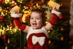 happy baby girl in red Santa dress arms raised by Christmas tree Christmas photoshoot Northampton